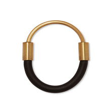 Load image into Gallery viewer, Bangle laying flat on white surface. This bangle is made from a curved brass rod made to fit halfway around the wearer&#39;s wrist. The ends of the rod is joined by sliding each end into a black rubber cord with brass attachments and enclose around the wrist