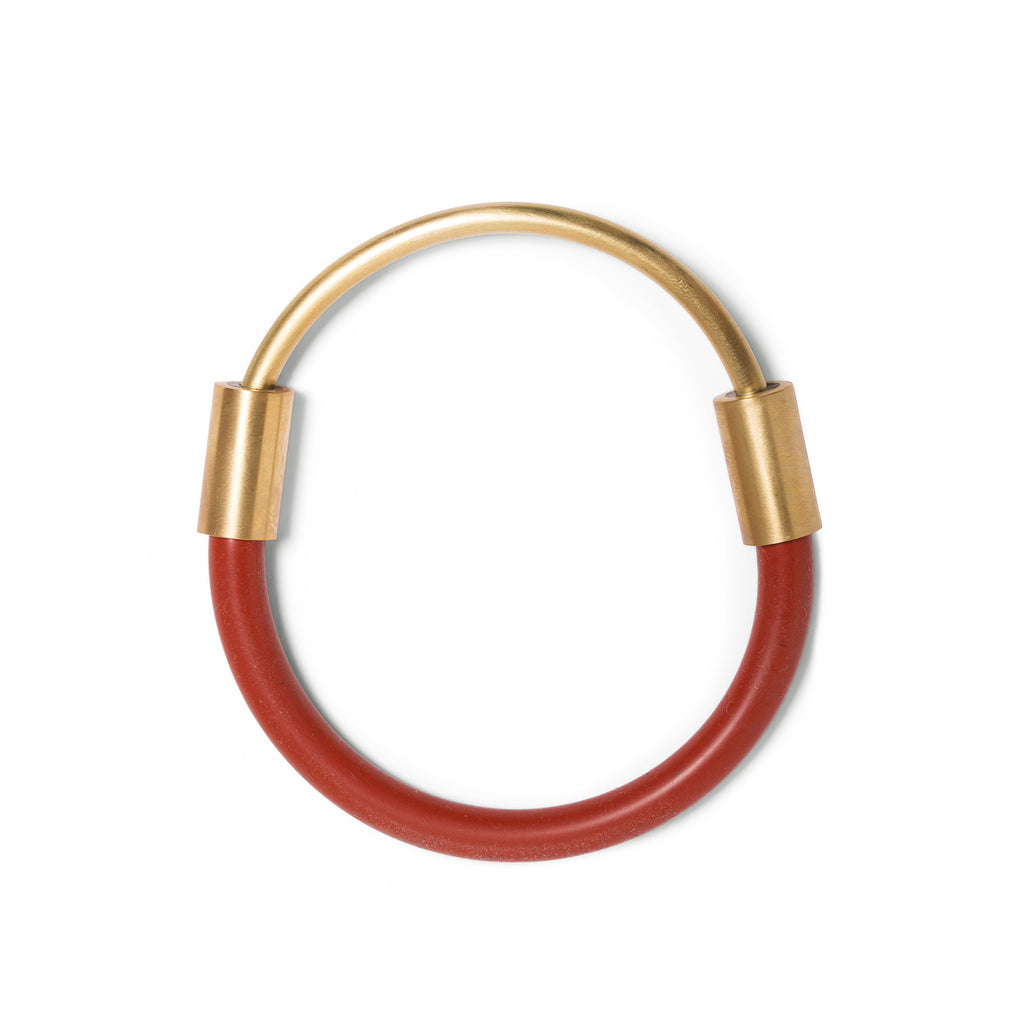 Bangle laying flat on white surface. This bangle is made from a curved brass rod made to fit halfway around the wearer's wrist. The ends of the rod is joined by sliding each end into a rusty-red rubber cord with brass attachments and enclose around the wrist