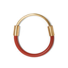 Load image into Gallery viewer, Bangle laying flat on white surface. This bangle is made from a curved brass rod made to fit halfway around the wearer&#39;s wrist. The ends of the rod is joined by sliding each end into a rusty-red rubber cord with brass attachments and enclose around the wrist