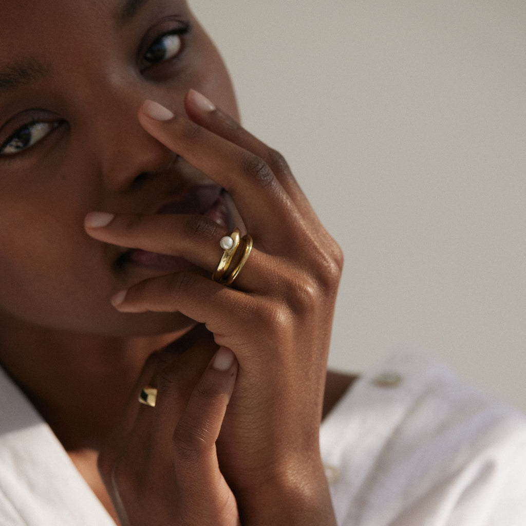 Black woman standing up-close to camera with hands close to face, looking into camera. Wearing gold pearl ring stacked on thinner, plain gold ring.