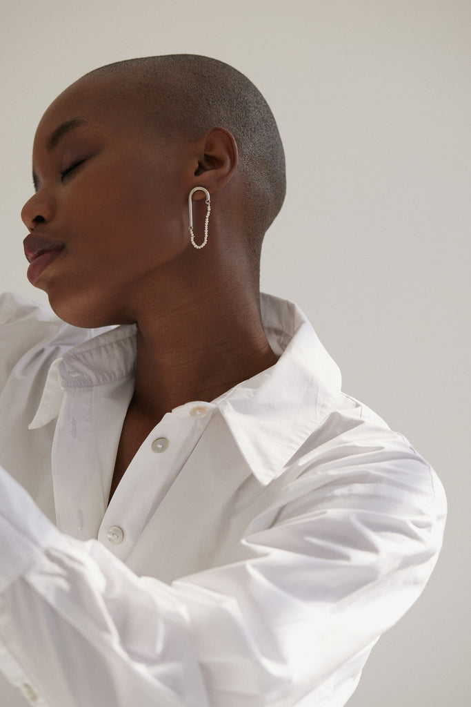 Black bald woman wearing white collar shirt turning to left with closed eyes looking relaxed. She's wearing earrings made from Silver in a cane shape with the two ends joined by a string of miniature pearls