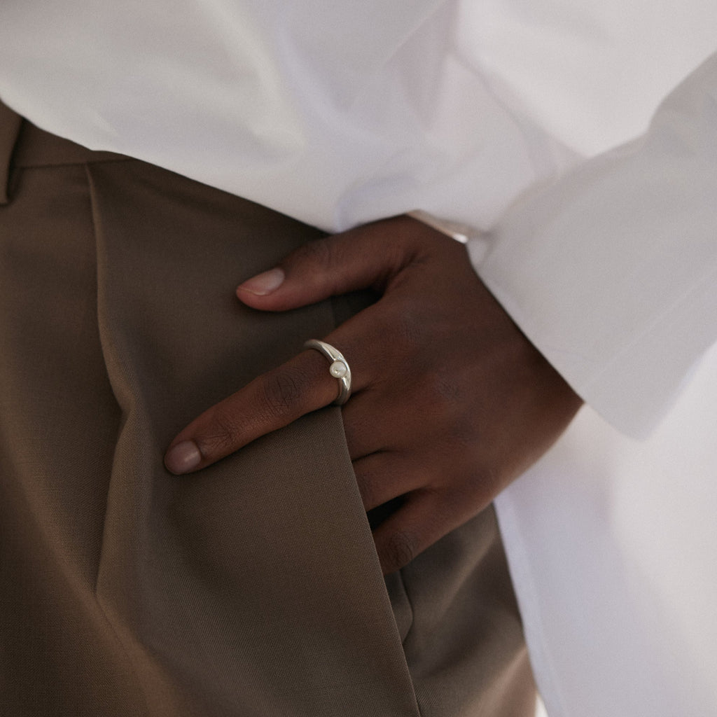 View of black woman's hand halfway in trouser pocket allowing Silver pearl ring to be visible on index finger. Wearing a white shirt and brown trousers